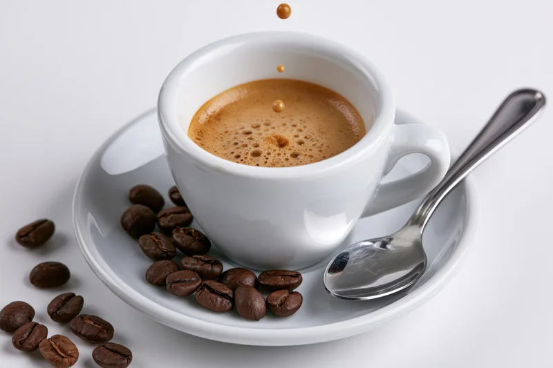 White ceramic espresso cup with foam and falling droplets on matching saucer, surrounded by coffee beans and a spoon, isolated on white background
