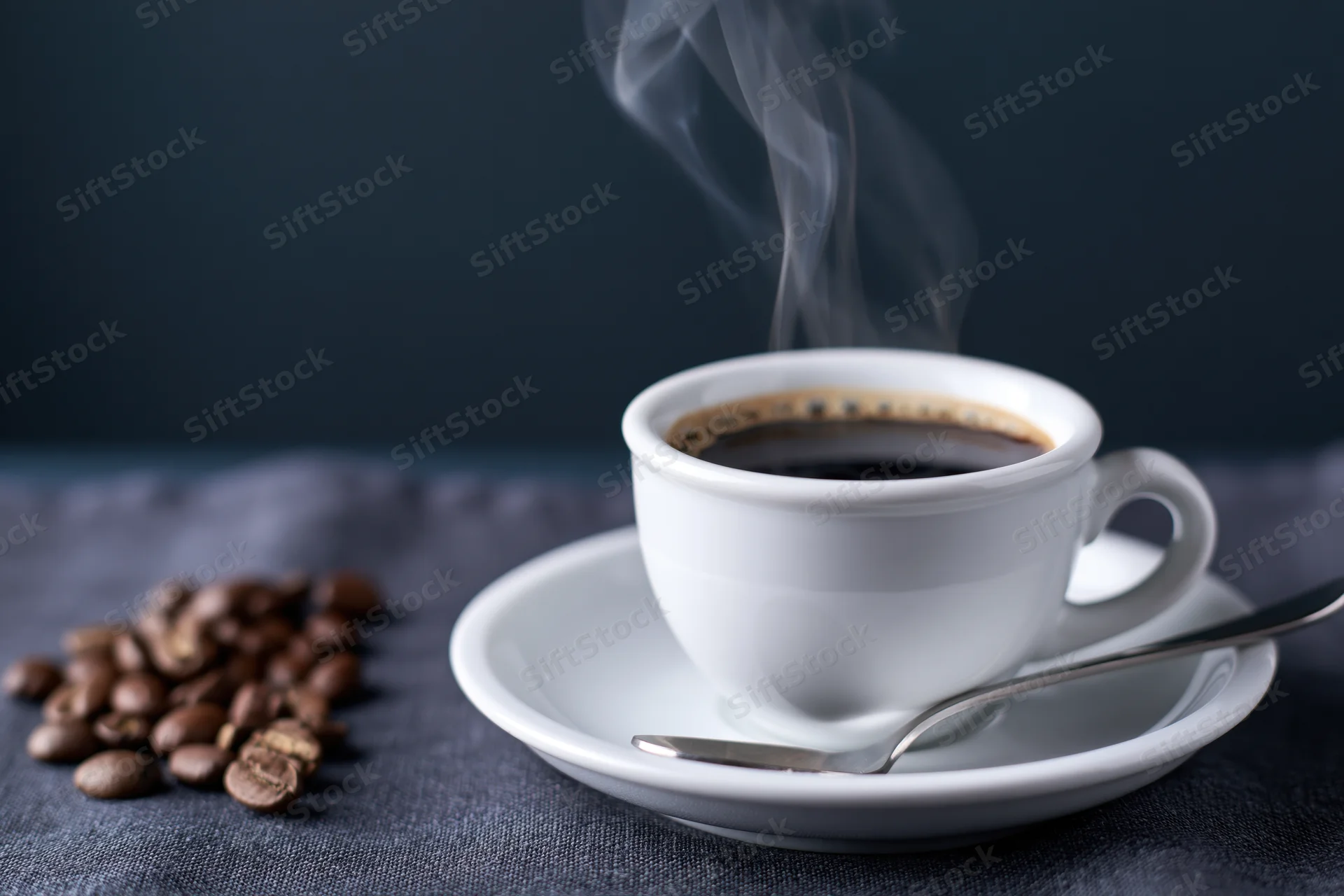 Steaming espresso in a white ceramic cup on a saucer with coffee beans beside it on a dark fabric surface