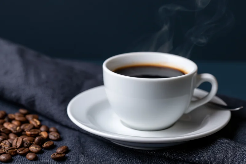 Steaming espresso in a white ceramic cup on a saucer with coffee beans beside it on dark fabric