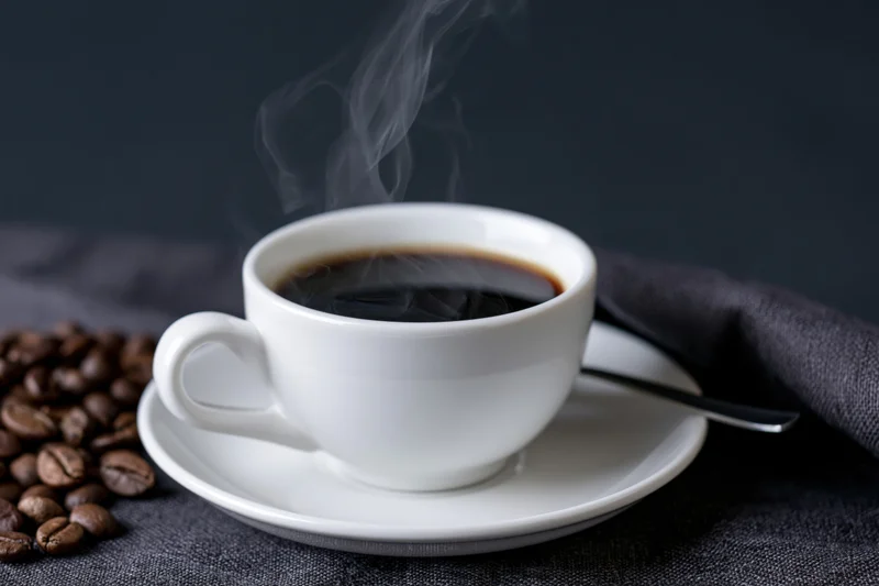 Steaming coffee in a white ceramic cup on a saucer with coffee beans nearby on a dark cloth surface