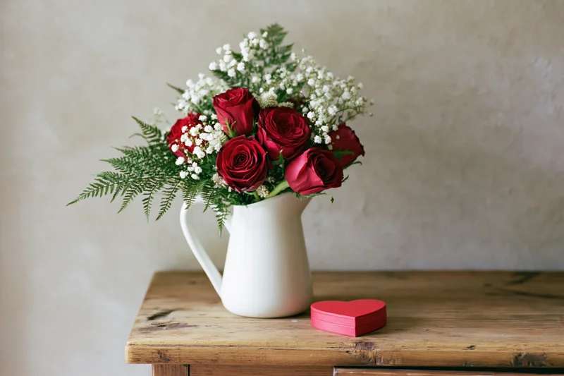 Red roses and white baby's breath in a white ceramic pitcher on a wooden table beside a red heart-shaped box