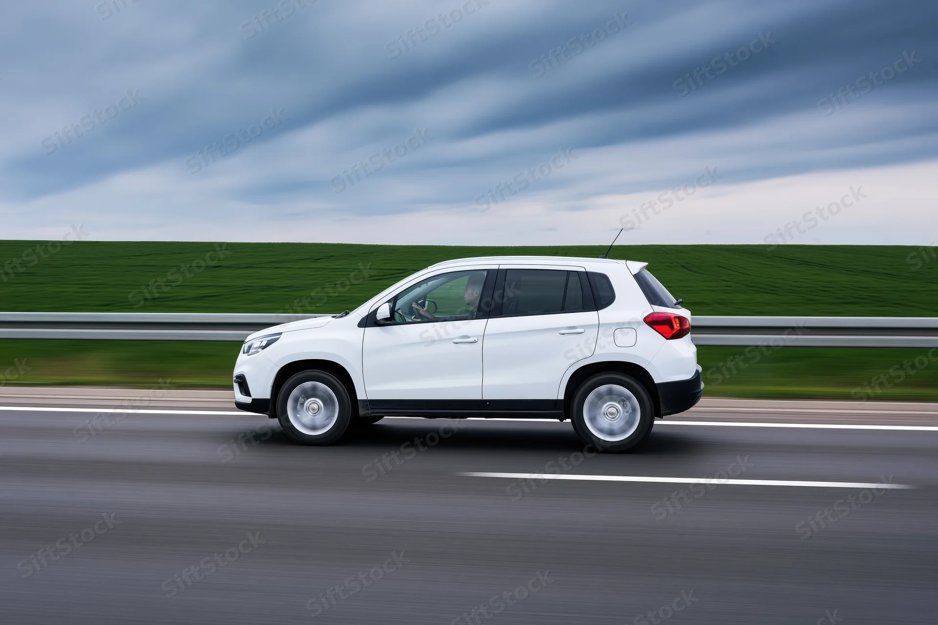 White compact suv driving on highway with blurred green field and cloudy sky in background