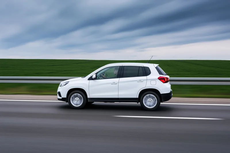 White compact suv driving on highway with blurred green field and cloudy sky in background