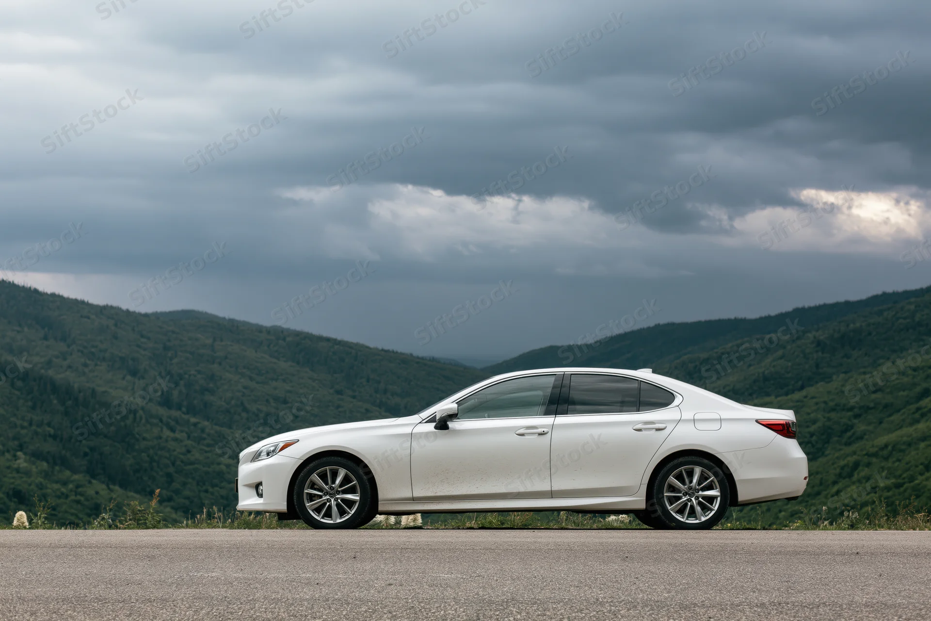 White sedan parked on mountain road shoulder with forested hills and cloudy sky in background