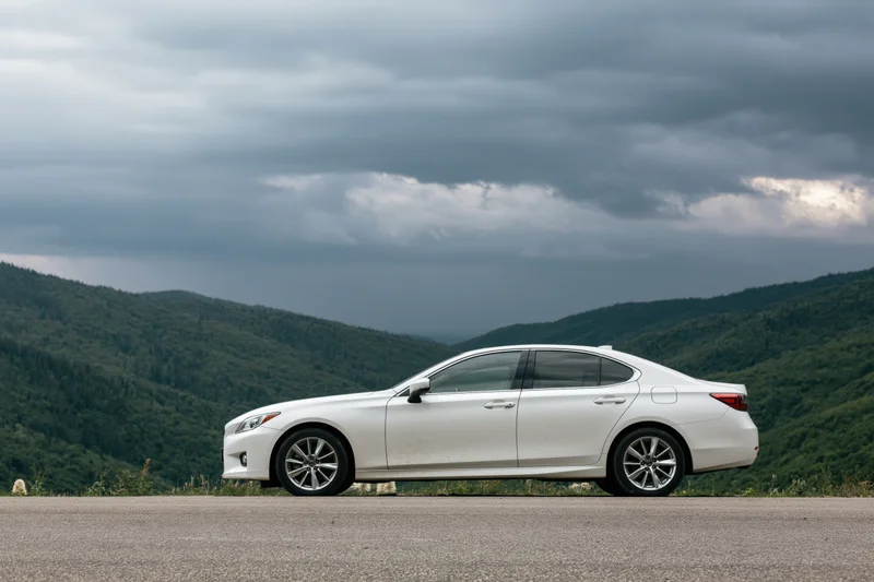 White sedan parked on mountain road shoulder with forested hills and cloudy sky in background