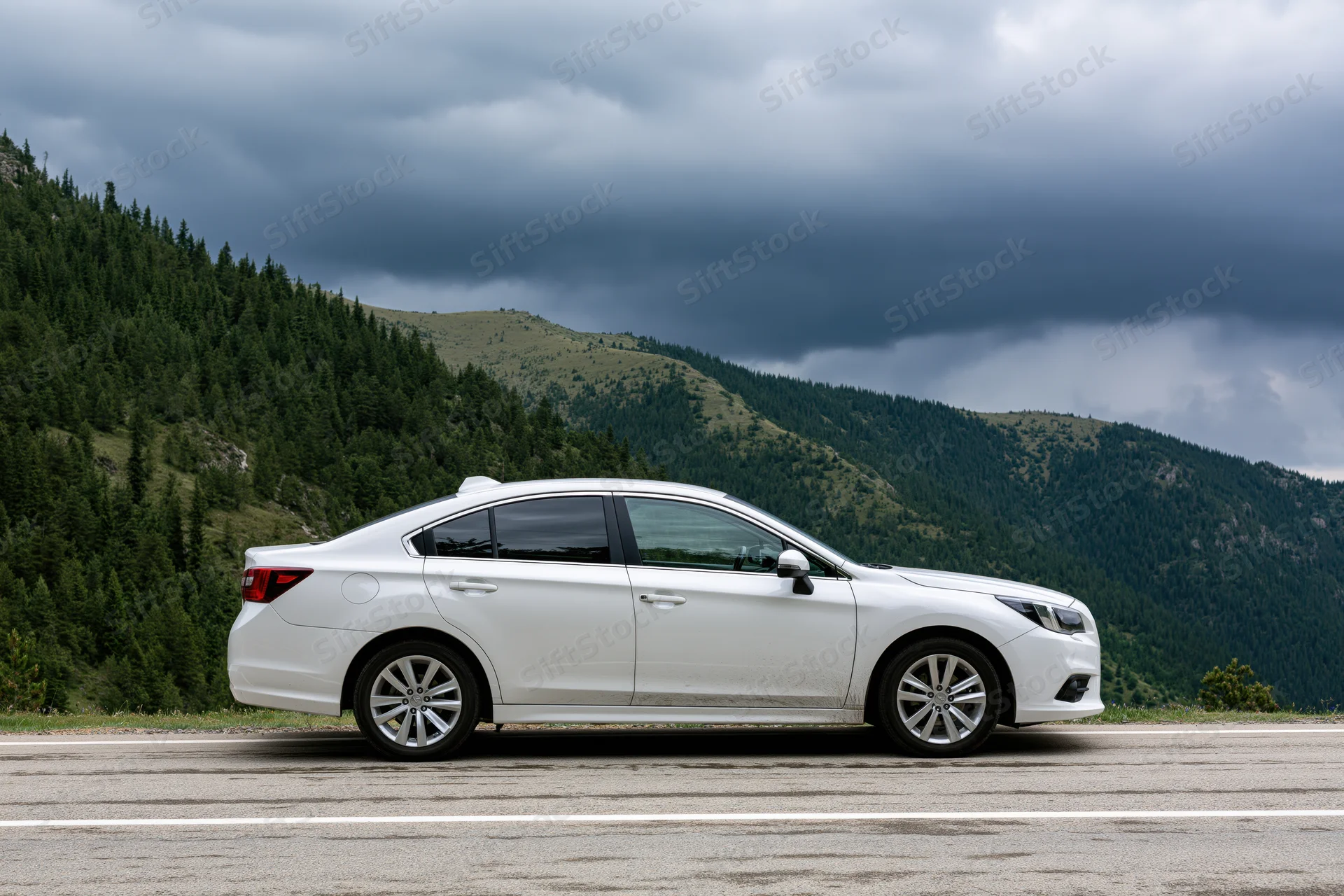 White sedan parked on mountain road shoulder with forested hills and cloudy sky in background