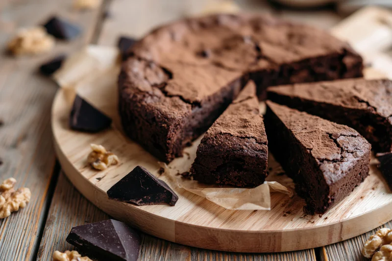 Sliced chocolate brownie cake on wooden board with chocolate chunks and walnuts