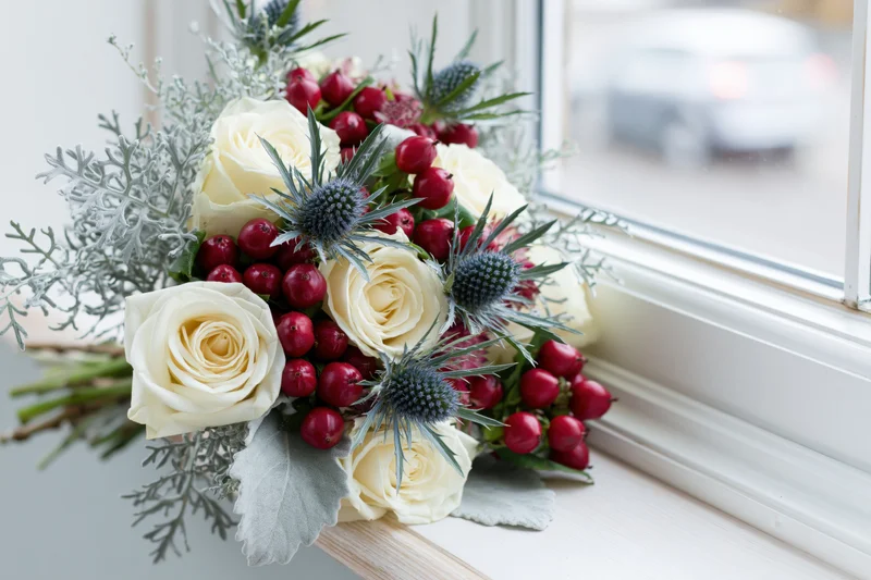 Winter bridal bouquet with white roses, red berries and thistle resting on a wooden windowsill