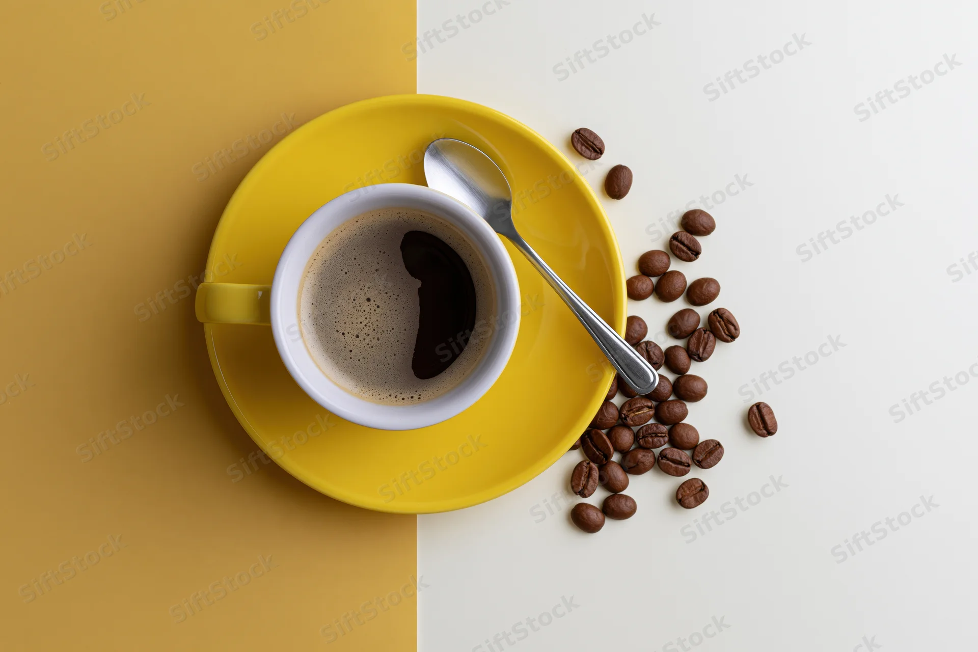 Yellow ceramic coffee cup with foam and scattered beans on a two-tone surface