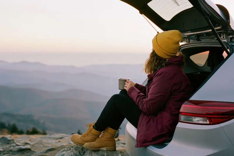 Woman sitting in open car trunk holding metal mug overlooking mountain range at sunset