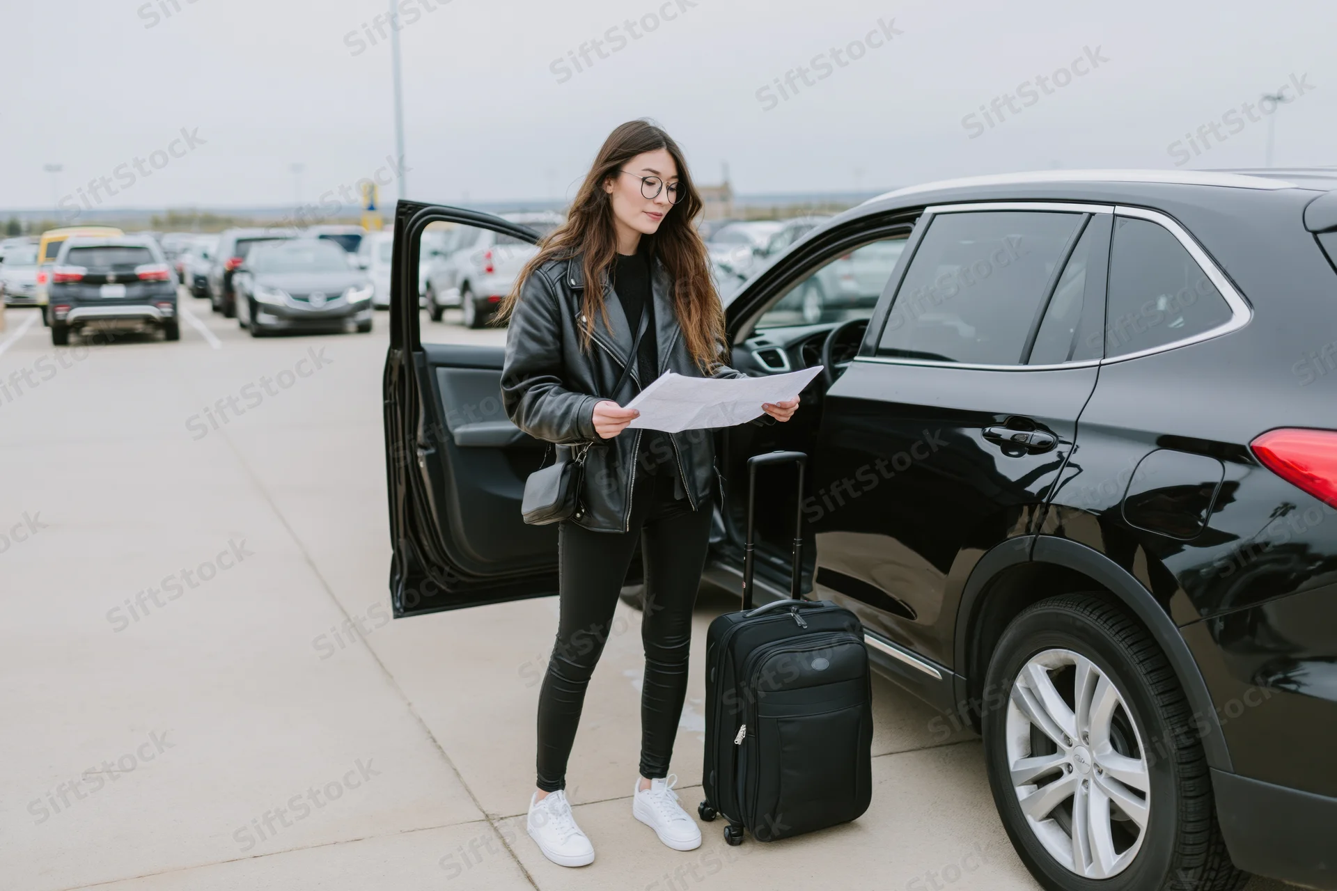 Woman reading a document beside an open car door with luggage in a parking lot