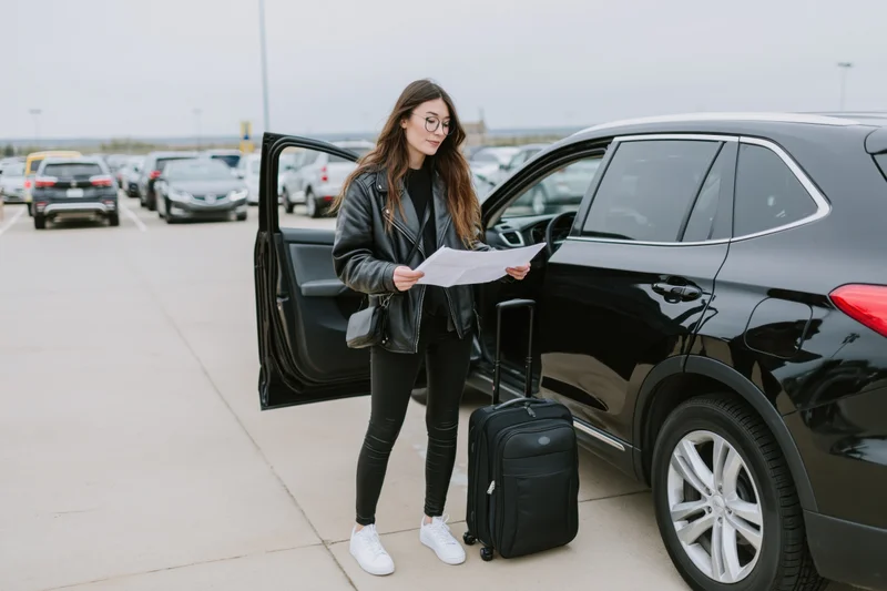 Woman reading a document beside an open car door with luggage in a parking lot