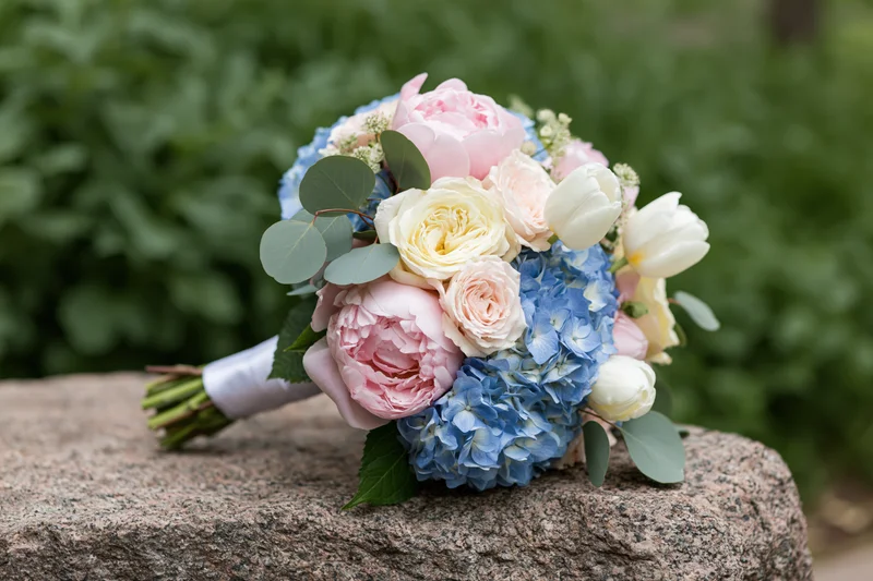 Bridal bouquet with pink peonies, blue hydrangeas and white tulips resting on a rough stone surface in a garden