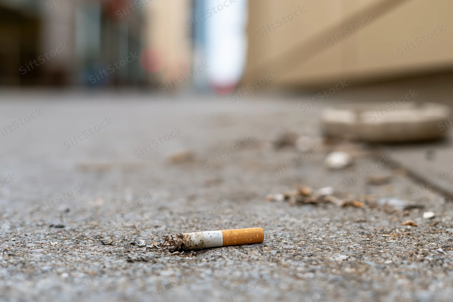 Cigarette butt lying on a concrete sidewalk with ash and litter around it