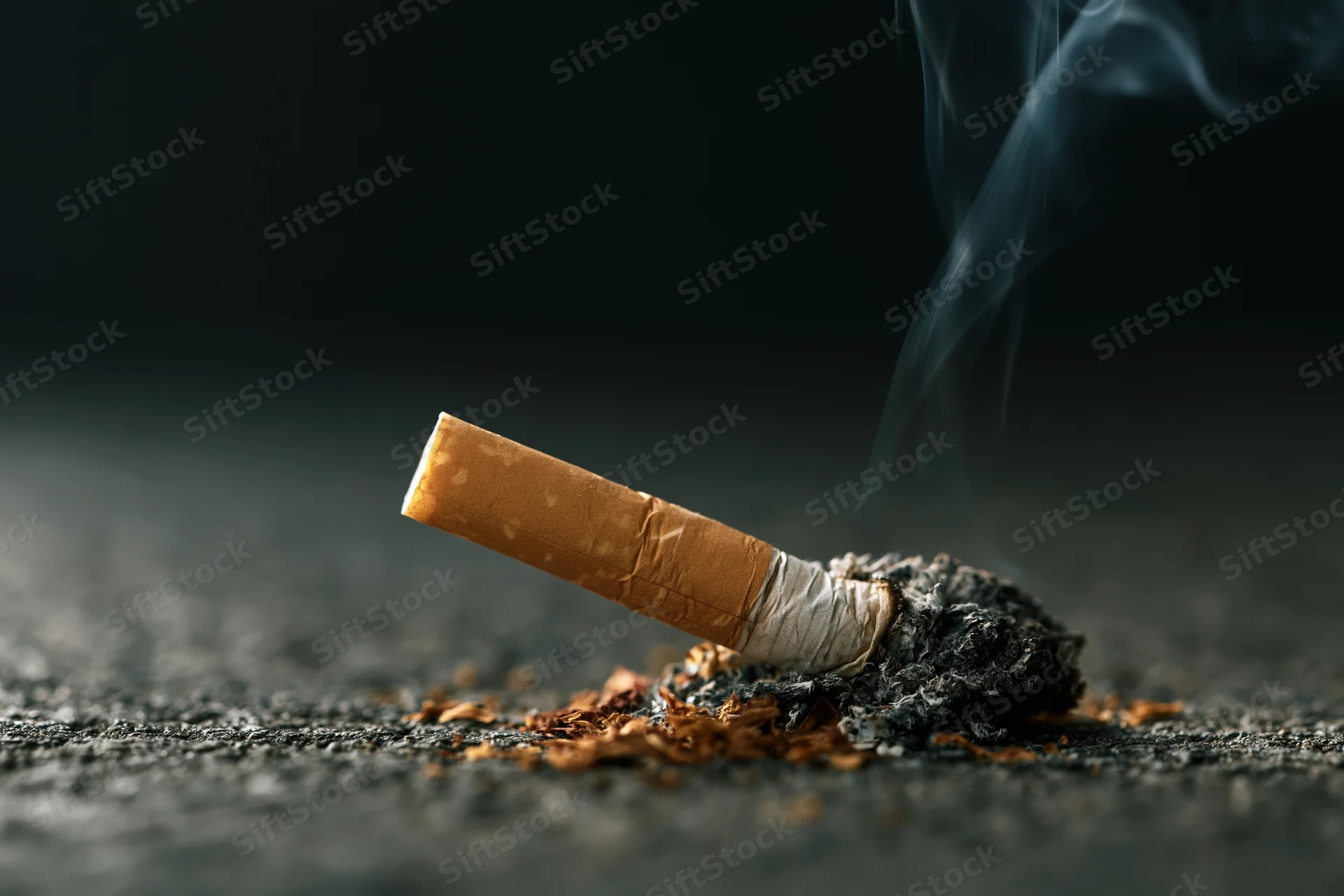 Cigarette butt with smoke rising from ash on dark concrete floor
