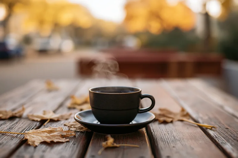 Steaming coffee cup on saucer with autumn leaves on weathered wooden table outdoors