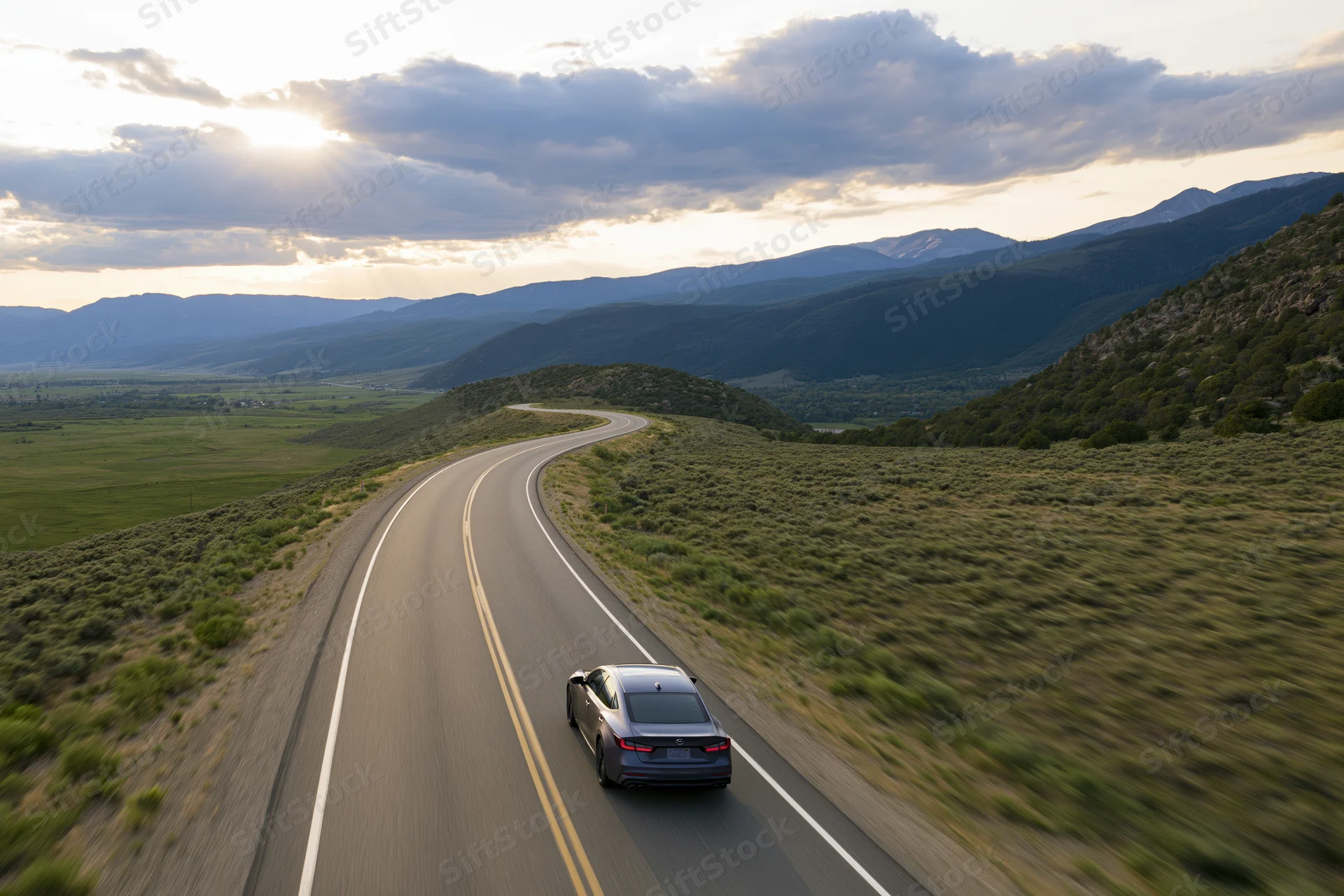 Dark sedan driving away on winding two-lane highway through mountainous rural landscape at sunset