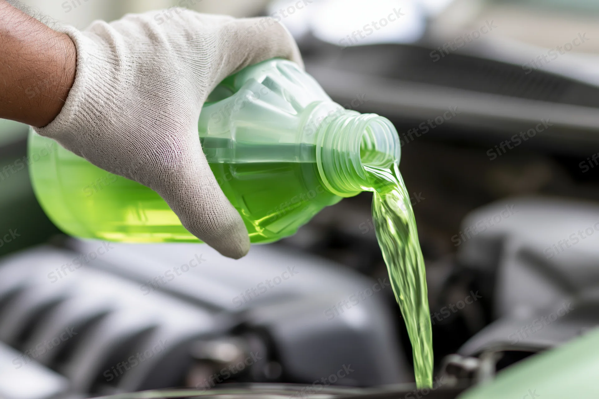 Gloved hand pouring green coolant into car engine from plastic bottle