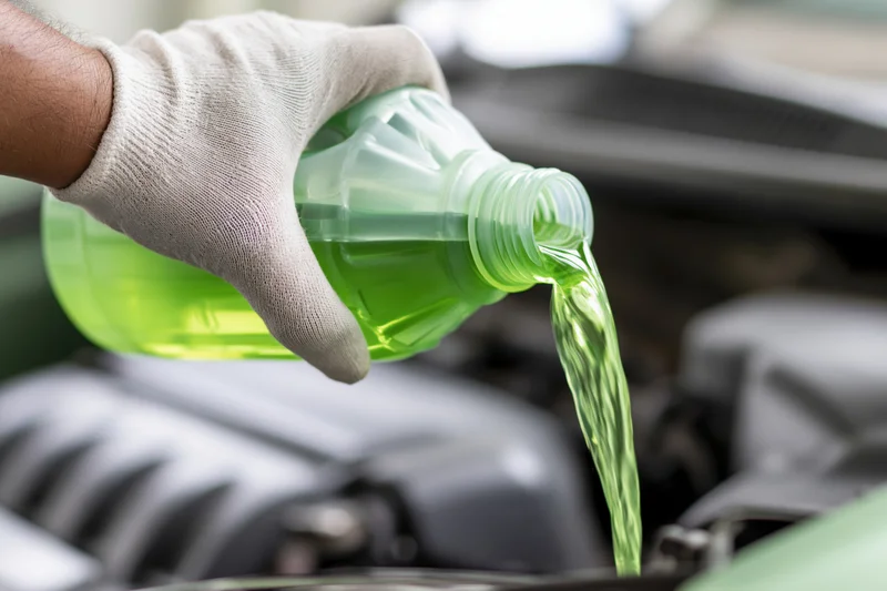 Gloved hand pouring green coolant into car engine from plastic bottle