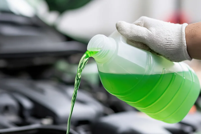 Gloved hand pouring green coolant into a car engine from a plastic bottle in a workshop