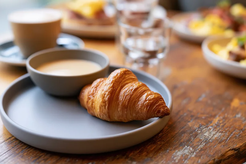 Golden flaky croissant on a matte grey ceramic plate with a bowl of cream on a wooden table