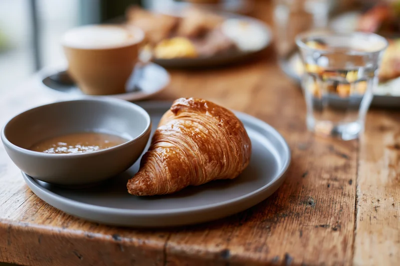 Golden flaky croissant on a matte grey ceramic plate with a bowl of soup on a wooden table in a cafe setting