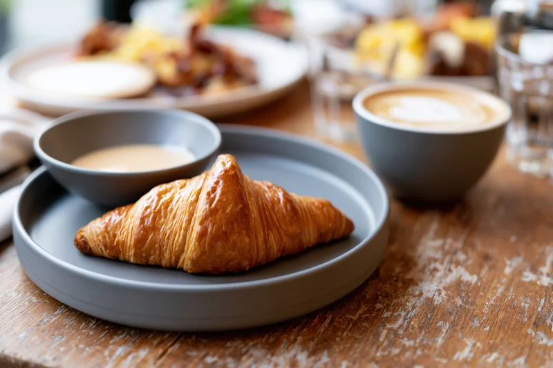 Golden flaky croissant on a matte grey ceramic plate with coffee and milk bowl on a wooden table