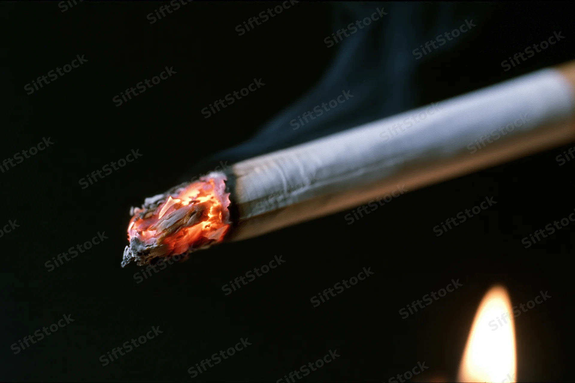 Lit cigarette burning with glowing ember and smoke against dark background, flame visible at lower right