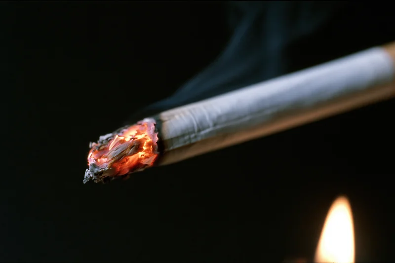 Lit cigarette burning with glowing ember and smoke against dark background, flame visible at lower right