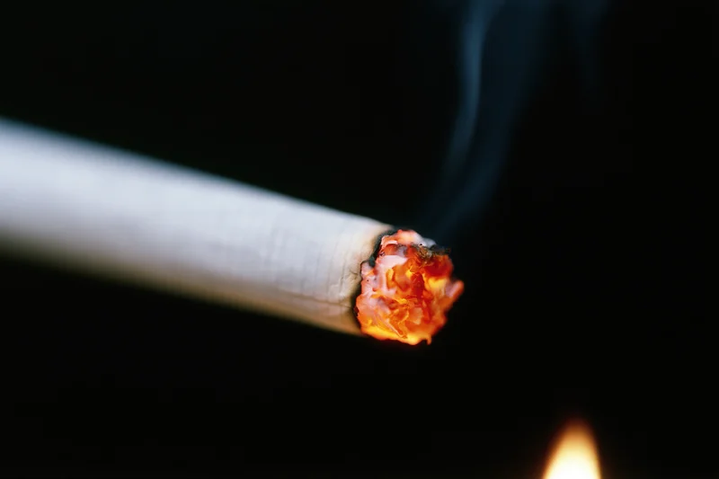 Lit cigarette burning with glowing ember and smoke against dark background, being ignited by a flame