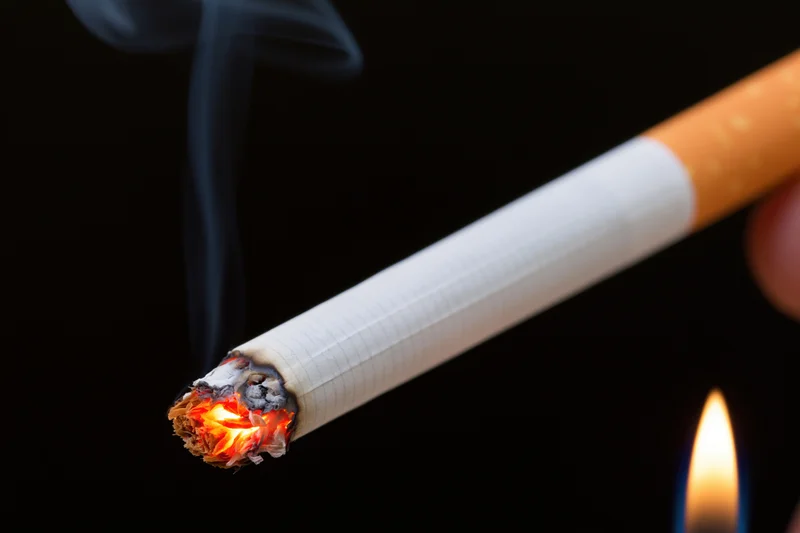 Lit cigarette held diagonally with glowing ember and rising smoke against black background