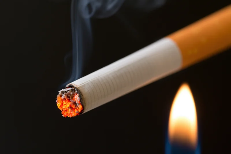 Lit cigarette with glowing ember and rising smoke against dark background, flame nearby