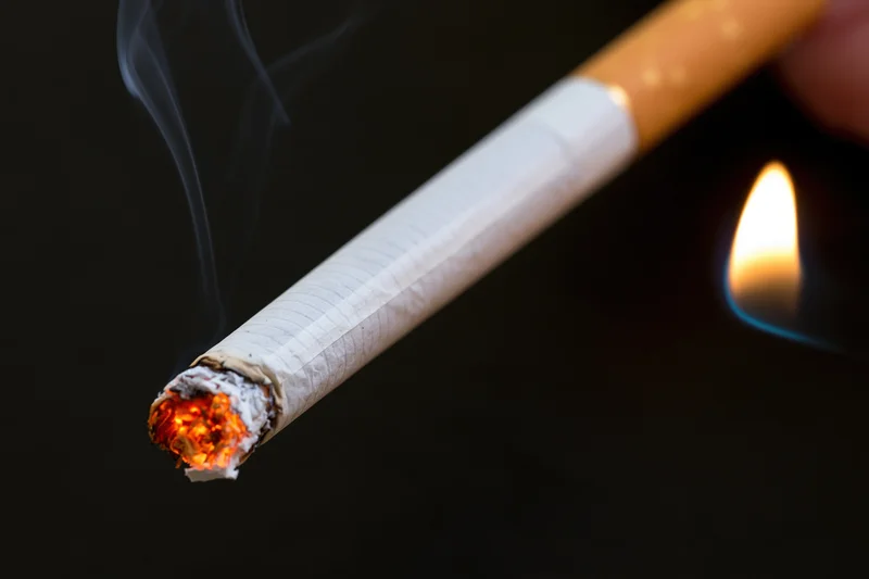 Lit cigarette with glowing ember and smoke against dark background, being lit by a flame