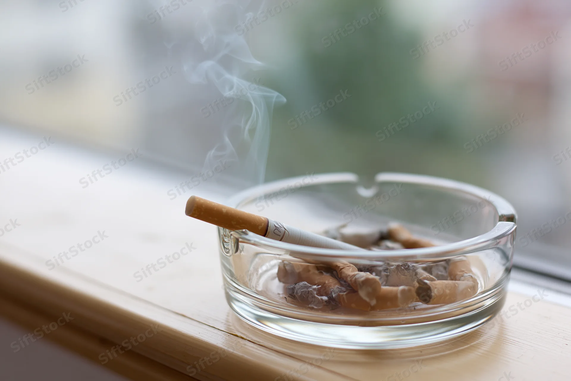 Lit cigarette in a glass ashtray on a wooden windowsill with smoke rising