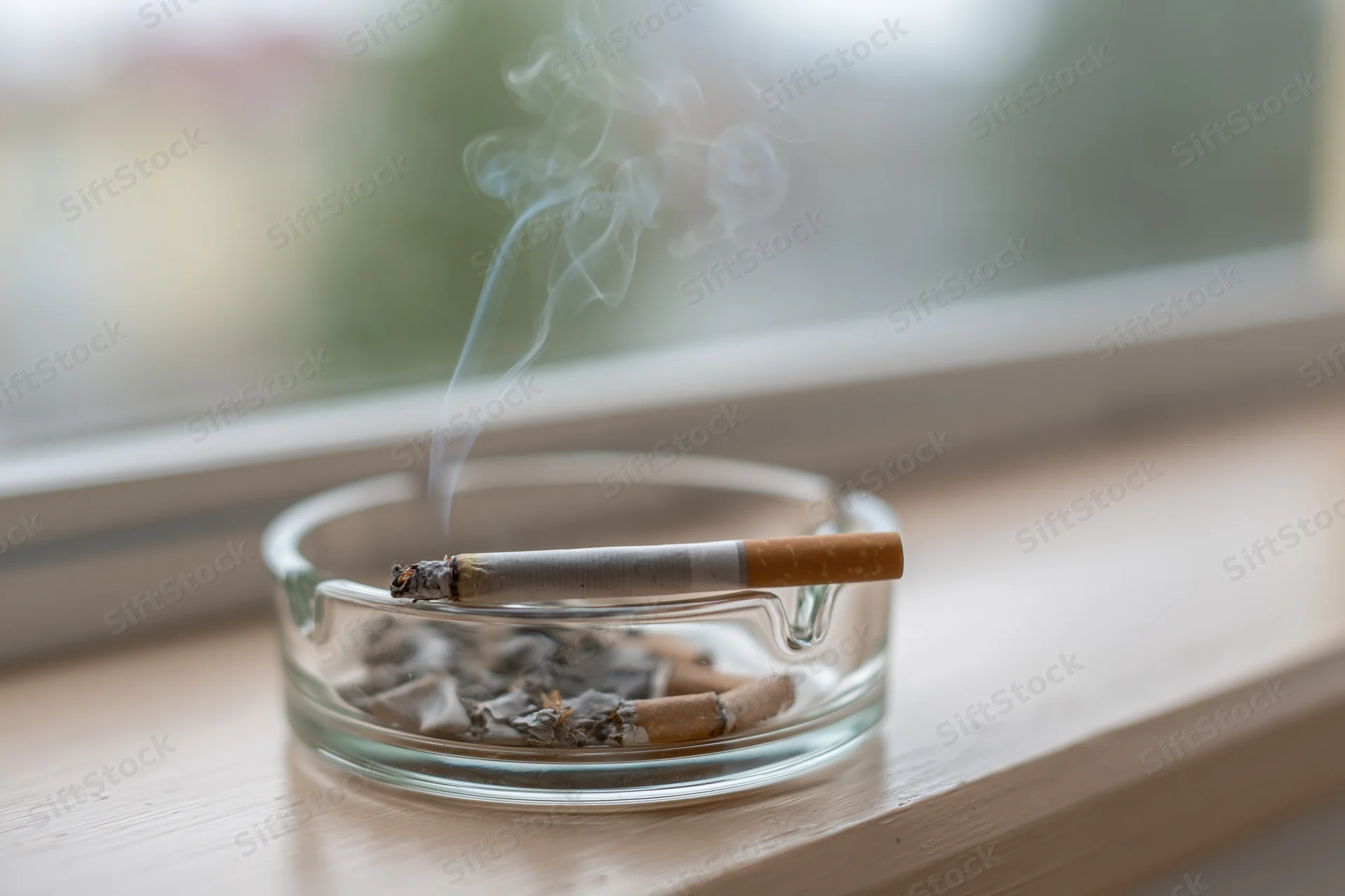 Lit cigarette burning in a clear glass ashtray on a wooden windowsill with smoke rising