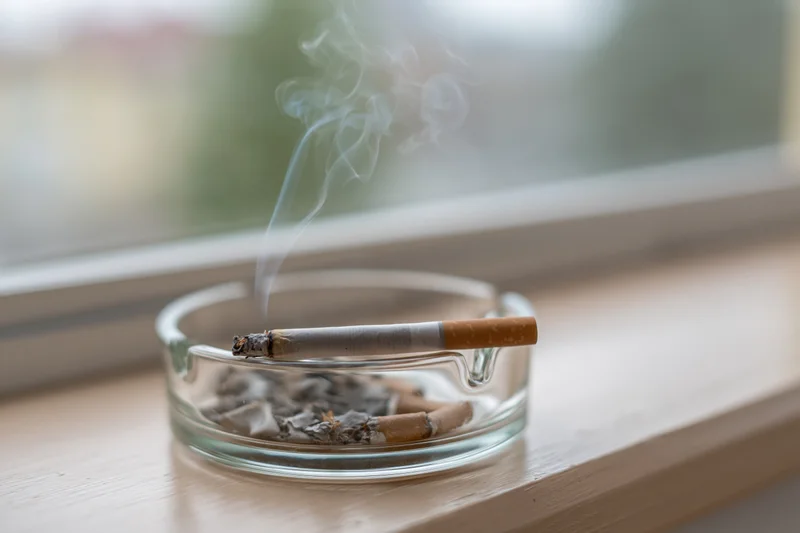 Lit cigarette burning in a clear glass ashtray on a wooden windowsill with smoke rising