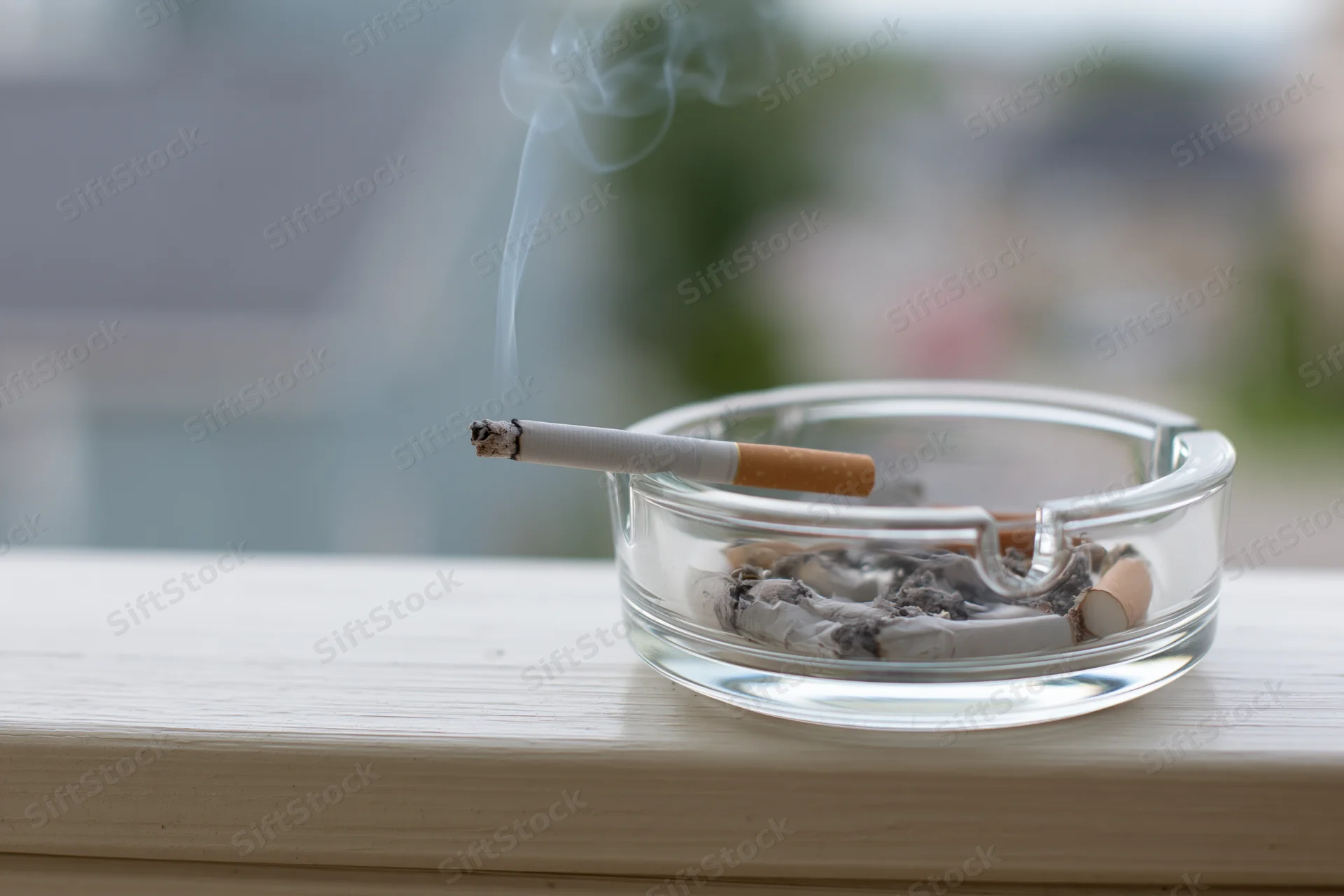 Lit cigarette resting in a clear glass ashtray on a wooden surface near a window