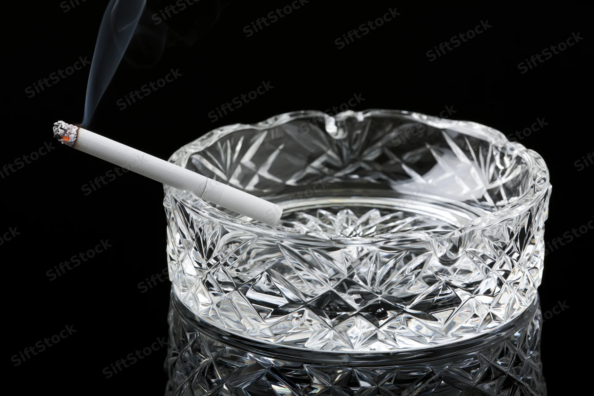 Lit cigarette resting in a heavy cut-crystal glass ashtray against black background