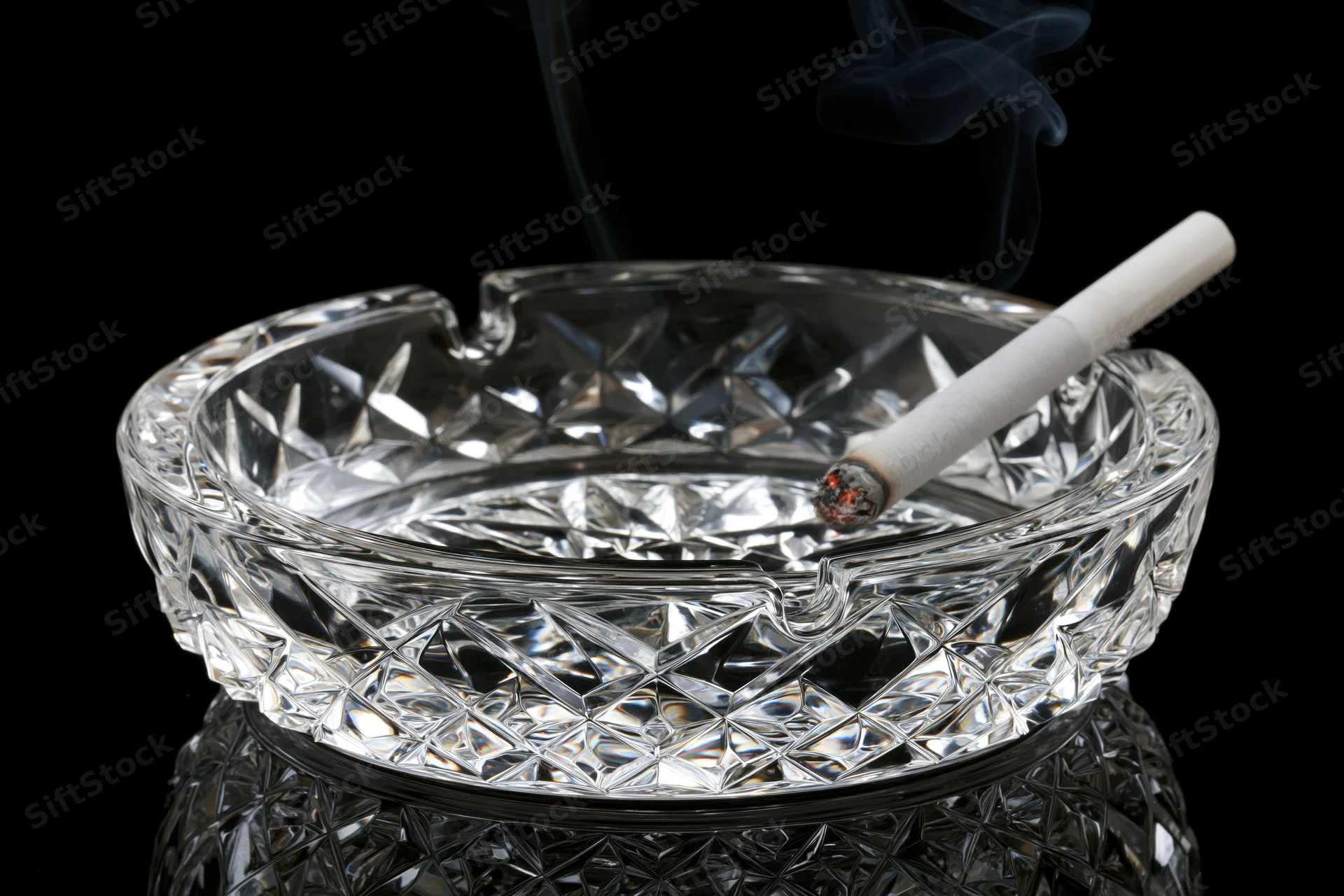 Lit cigarette resting in a heavy cut-crystal glass ashtray on black background