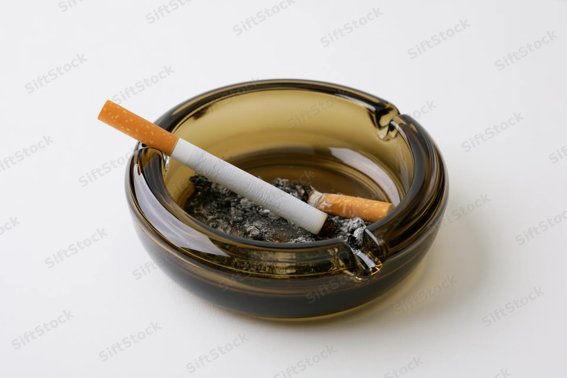 Lit cigarette resting in notch of dark glass ashtray filled with ash on white background