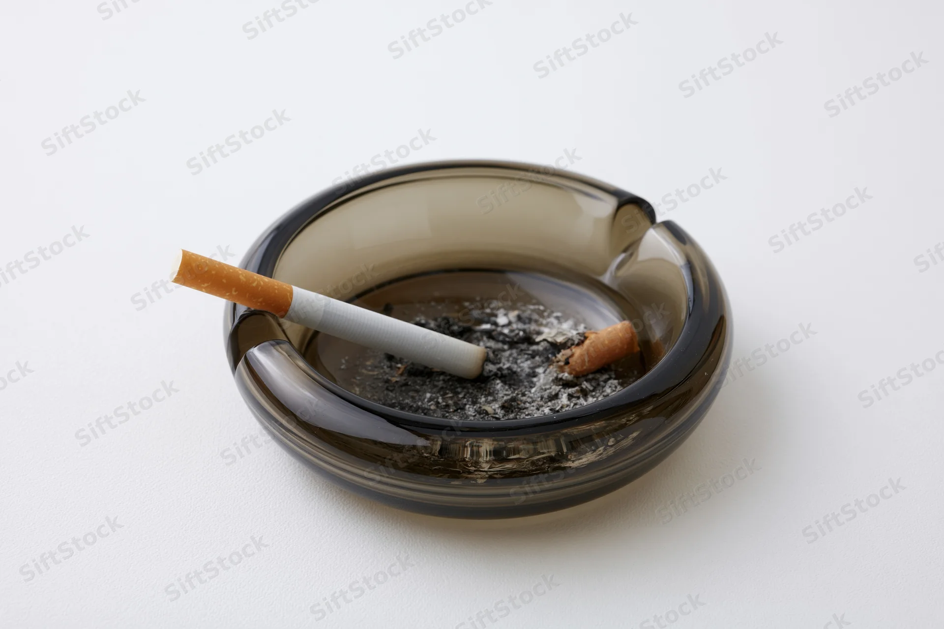 A cigarette resting in a dark glass ashtray filled with ash on a white surface