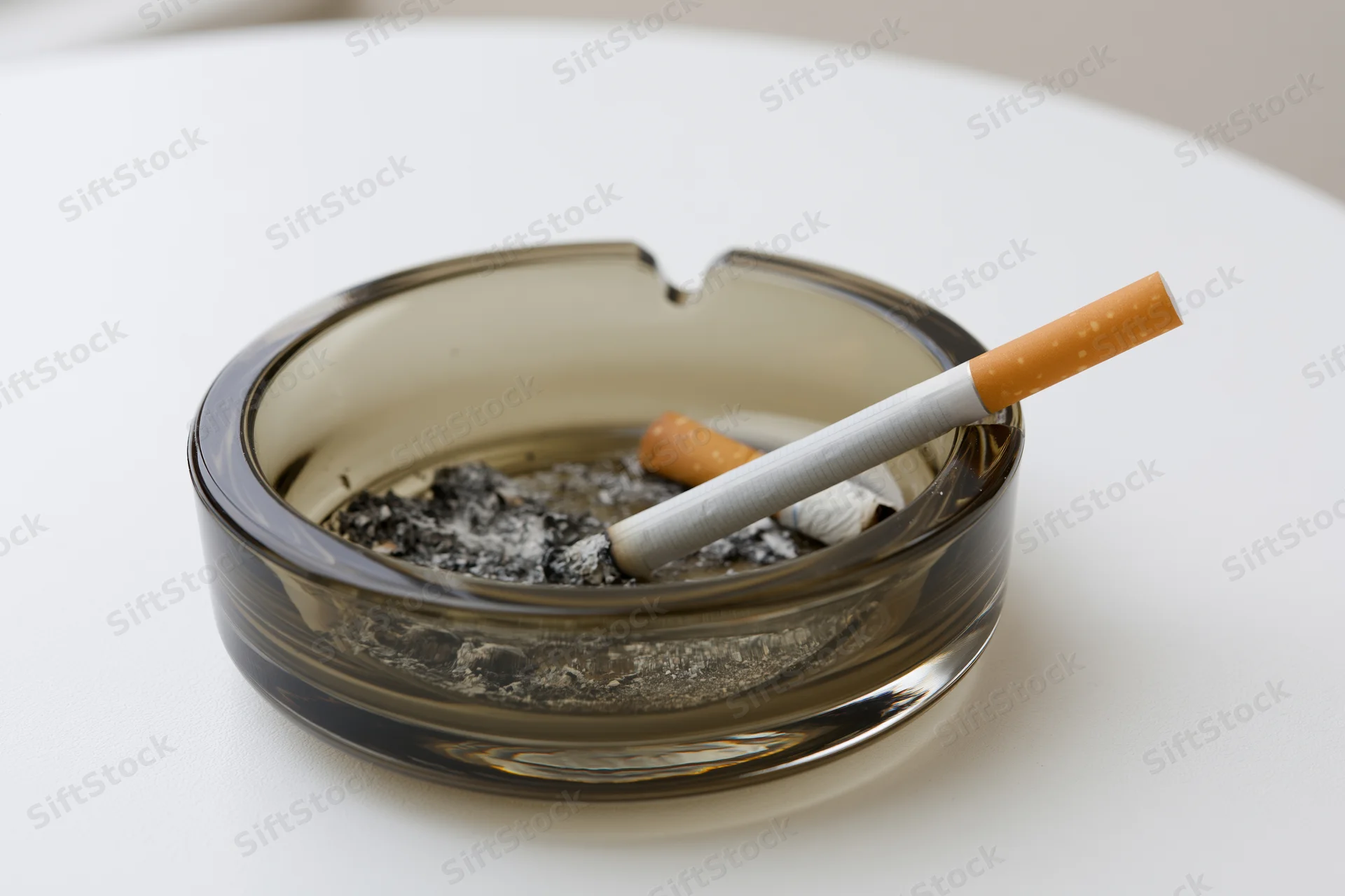 Lit cigarette resting in notch of dark glass ashtray on white table