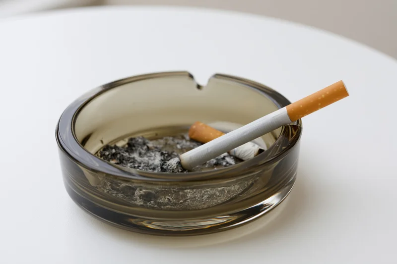 Lit cigarette resting in notch of dark glass ashtray on white table