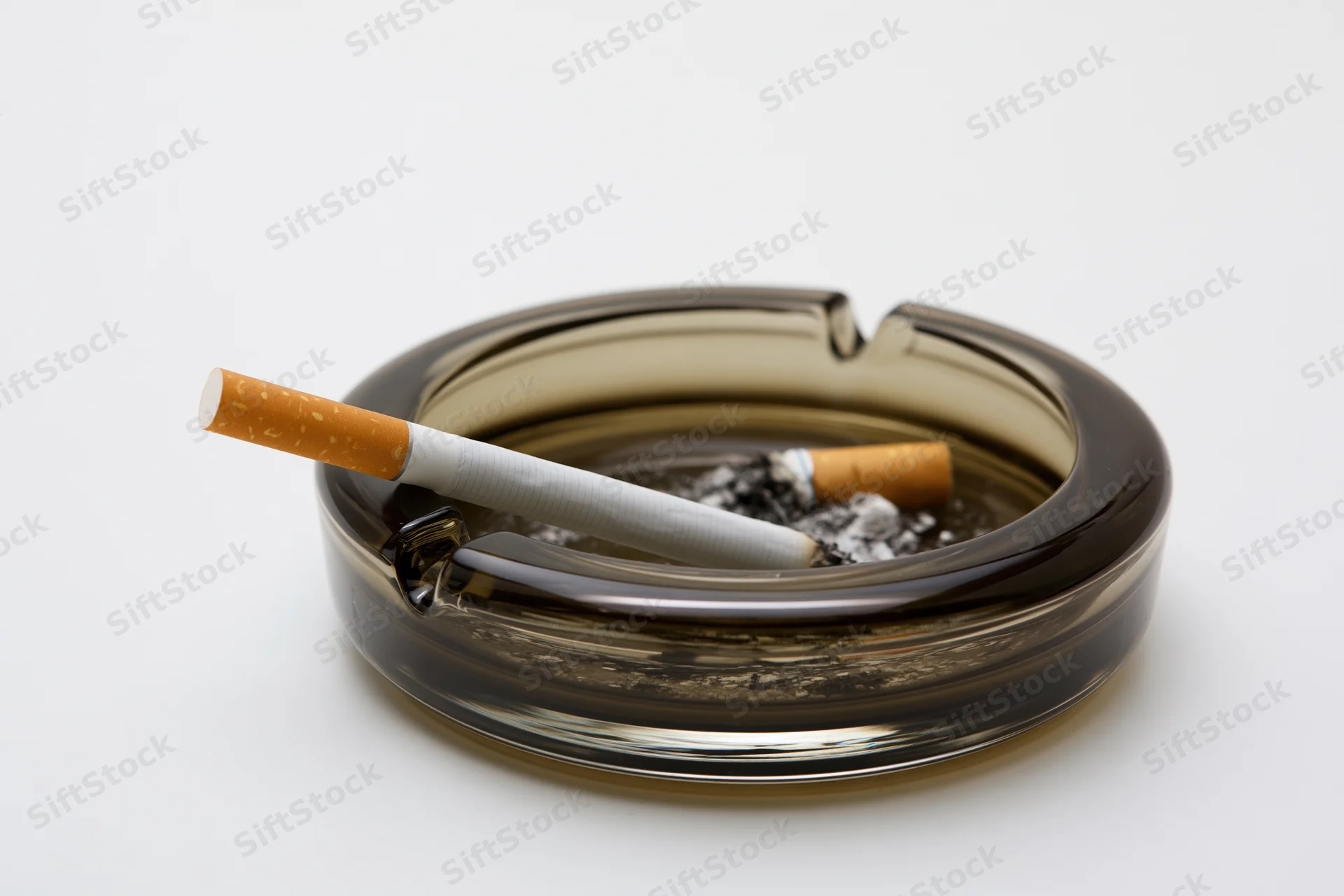 Lit cigarette resting in notch of dark smoky glass ashtray on white background