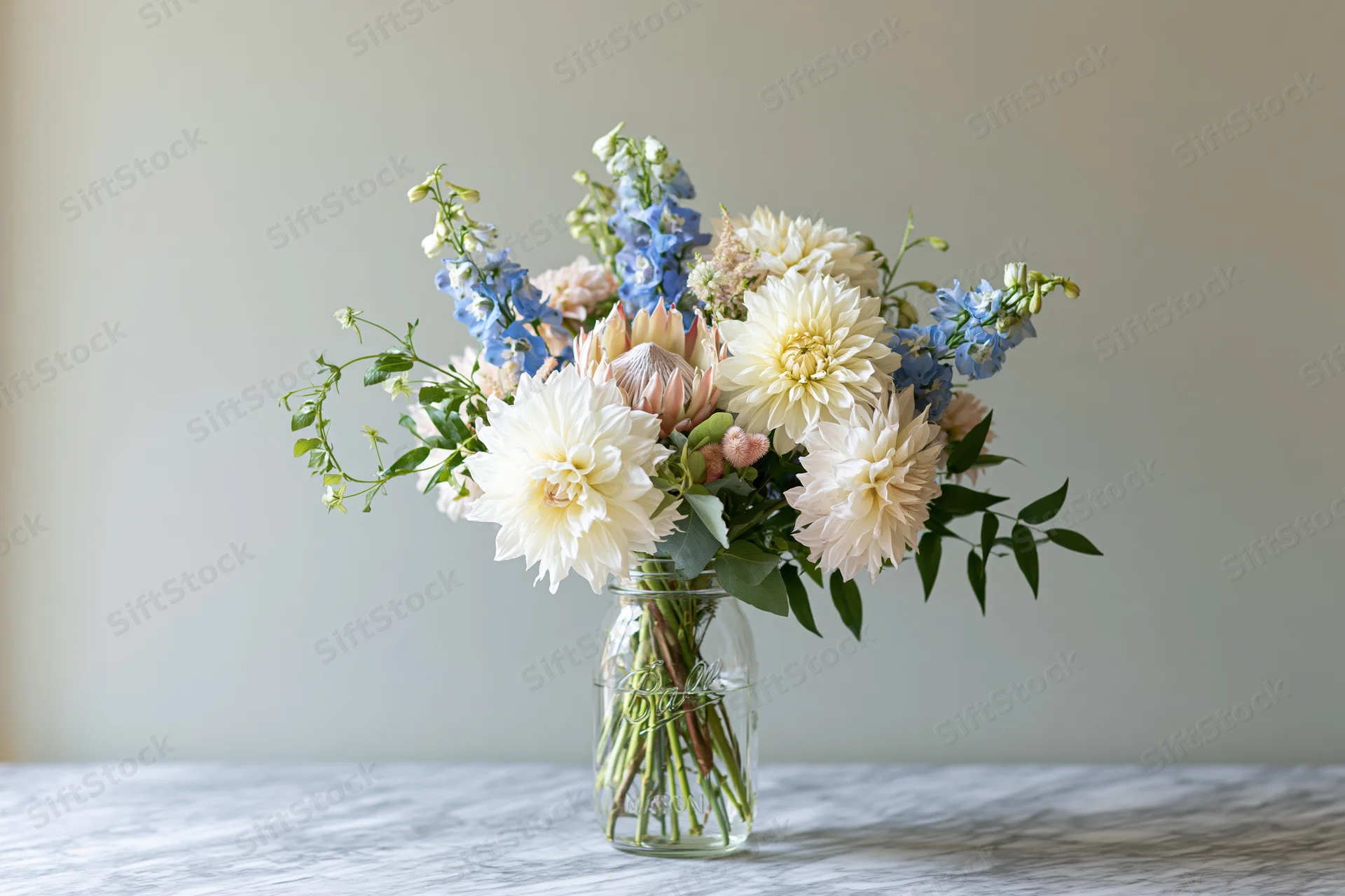 Loose garden bouquet of dahlias and delphiniums in a clear glass mason jar vase on a marble table