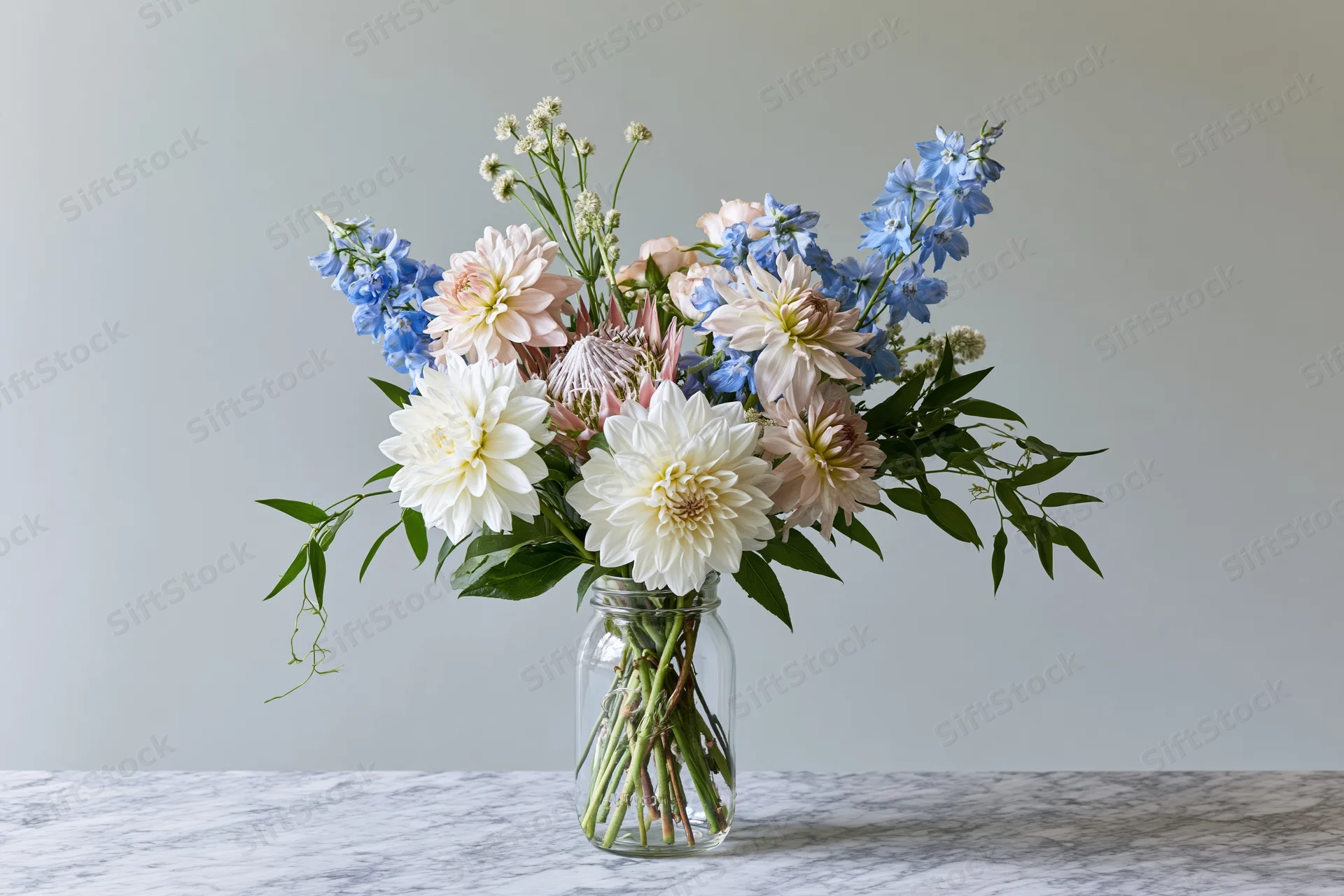 Loose garden bouquet of dahlias and delphiniums in a clear glass jar on a marble table