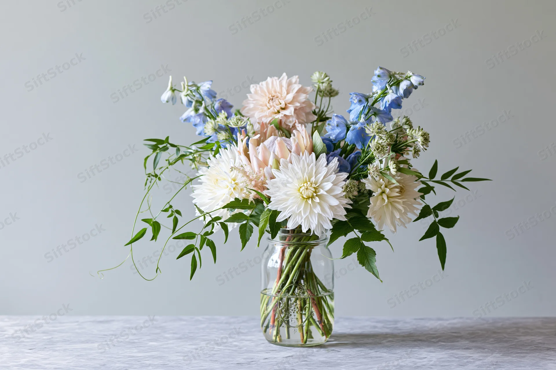 Loose garden bouquet of dahlias and clematis in a clear glass jar on a light surface