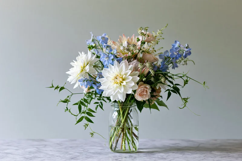Loose garden bouquet of dahlias, delphiniums, and roses in a clear glass jar vase on a light surface