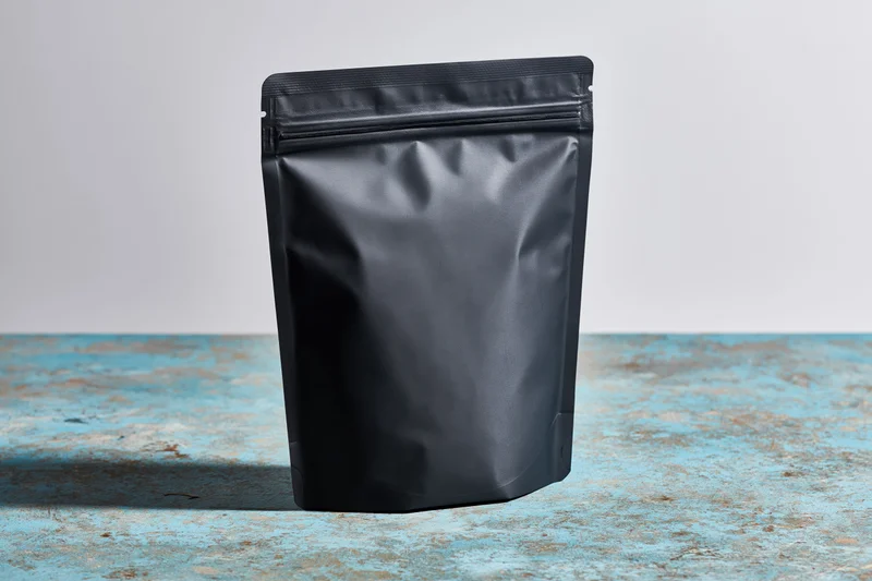 Matte black flat-bottom coffee pouch with zip seal standing on blue textured surface
