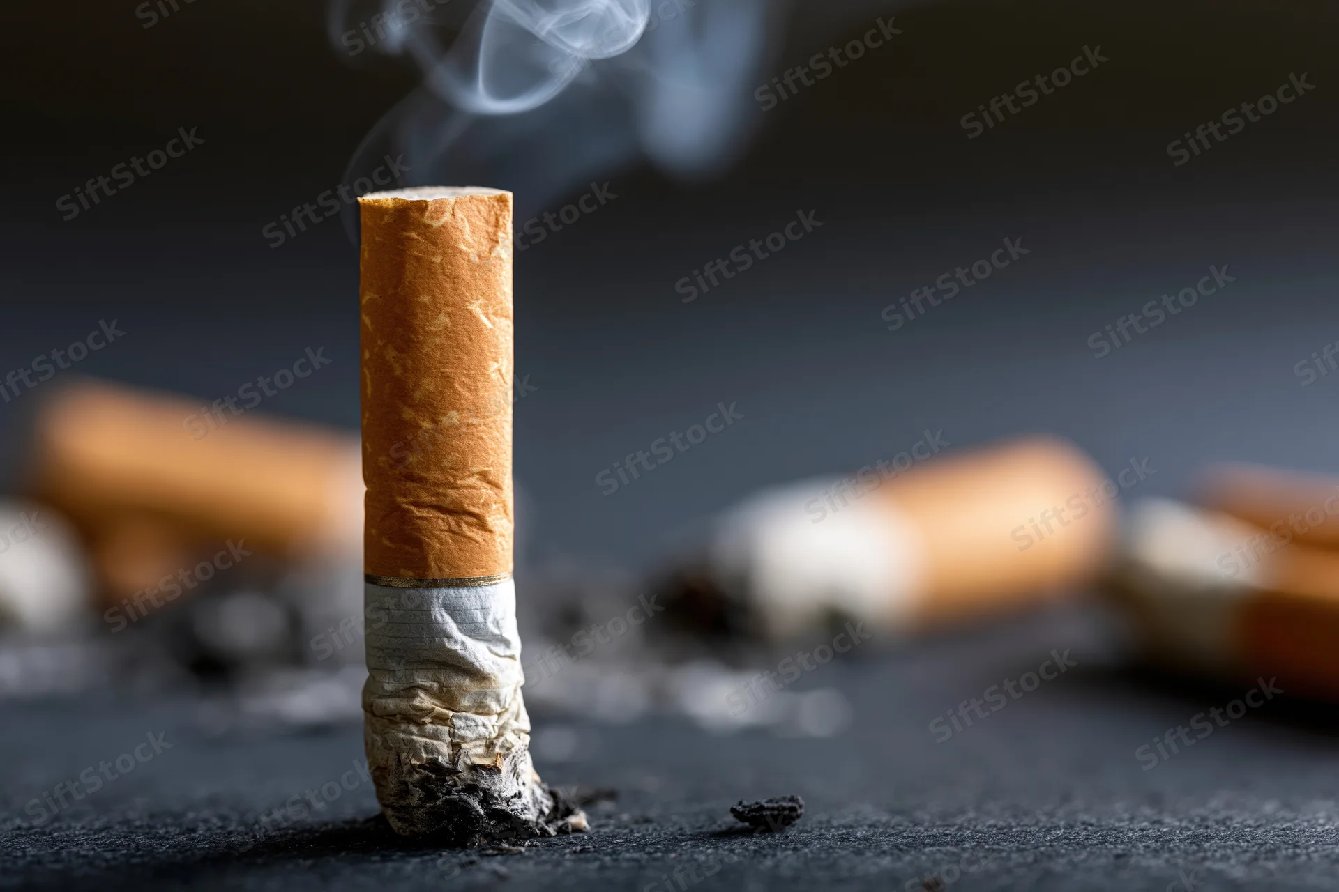 A smoking cigarette butt standing upright on a dark textured surface with smoke rising and ash scattered around